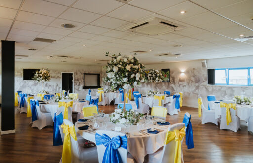 event space, nicely decorated, fine dining, yellow and blue ribbons behind white covered chairs, flower centrepieces